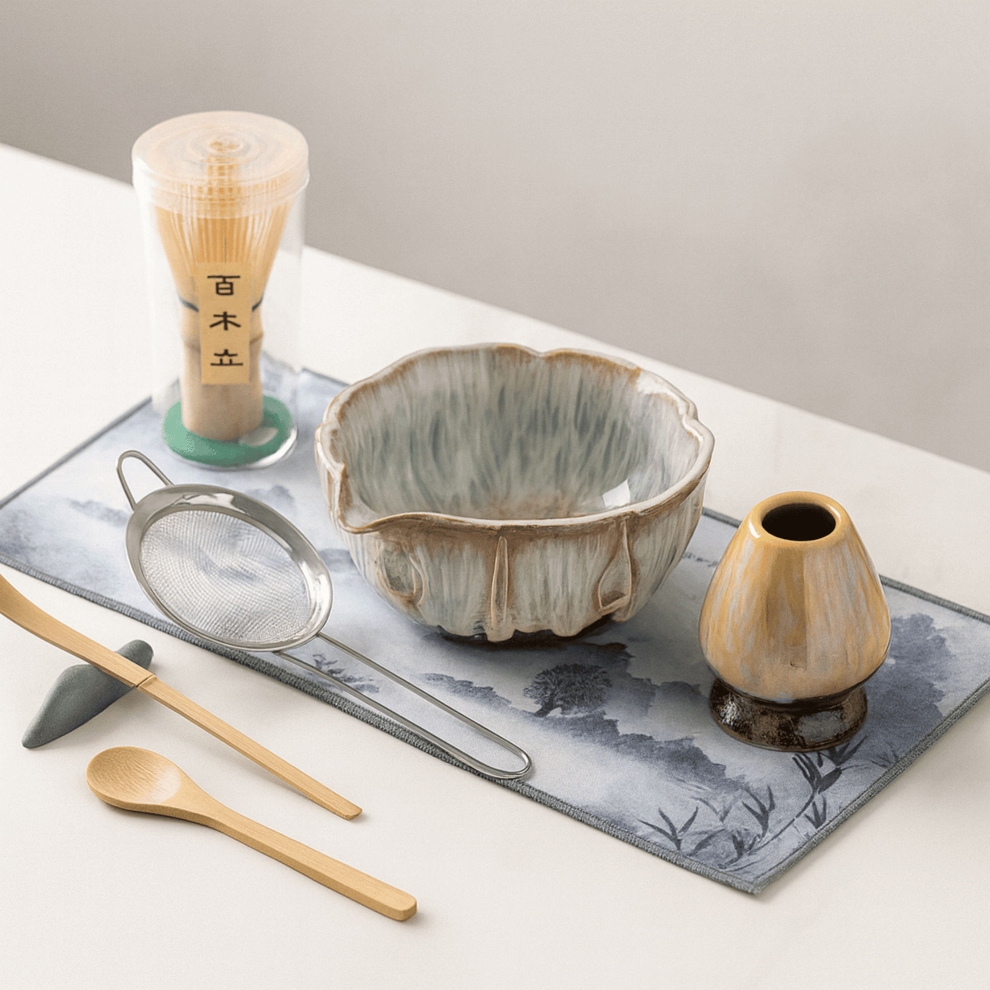 Tea-making set with ceramic bowl, whisk, spoon, and small vase on a decorative mat.