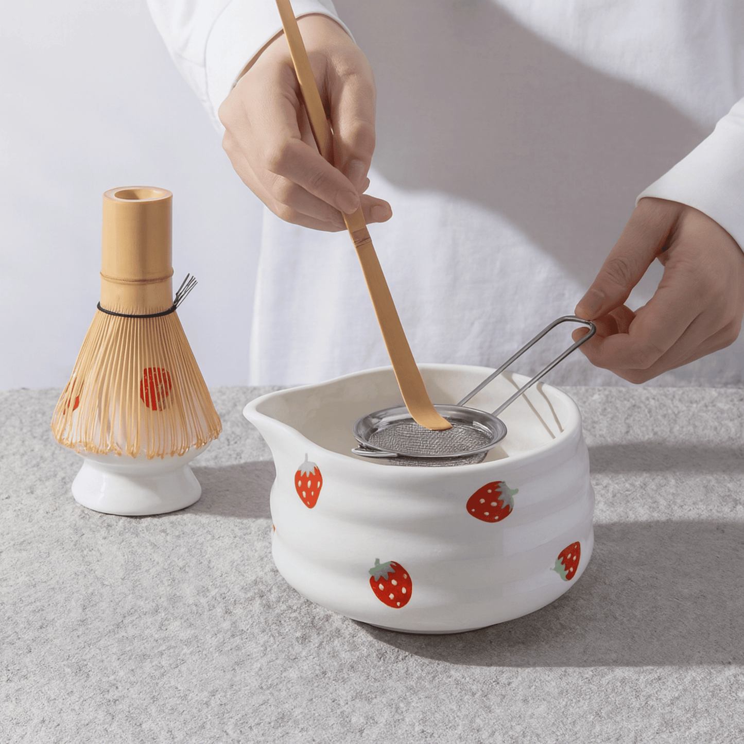 Person using a wooden whisk and strainer over a white ceramic pot with strawberry design on a light gray background.