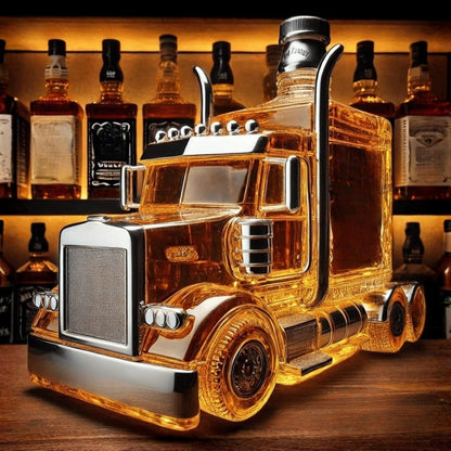 A large, glass semi-truck shaped whiskey decanter filled with amber bourbon, sitting on a wooden bar top with a shelf of liquor bottles in the background.