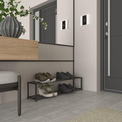 A minimalist black metal shoe rack positioned in a sleek, modern entryway. The rack features two tiers, holding various shoes, including black formal shoes and casual sneakers. The stylish space includes a wooden bench with a green plant in a vase, a dark door with a digital lock, and a mirror reflecting the contemporary design.
